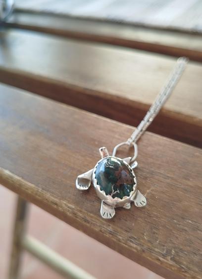 a pendant in shape of a turtle. it's silver with oval green gemstone. It's placed on a piece of wood