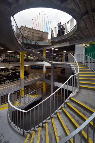 A spiral staircase in a grey, concrete carpark. At the top you can see out to the sky, and in the background is Edinburgh Castle with the Red Arrows flying over.