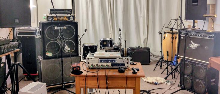 Photo of a rehearsal room. One bass amp, one guitar amp, both miced, on two sides of the table with recording equipment