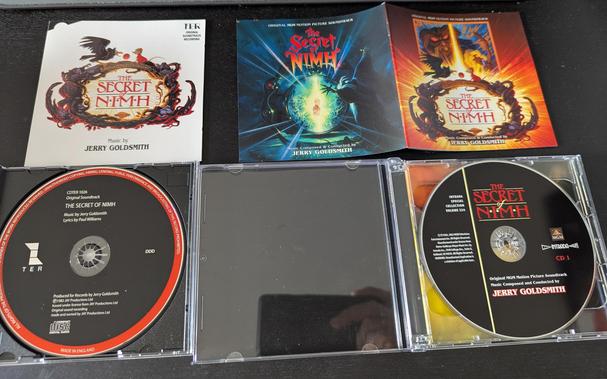 The old and the next disks and booklets next to each other.

The old cover shows Mes Brisby, a small, brown mouse with a red cape, and Jeremy the crow, on top of a big wooden frame made out of tree roots that encircle the text logo "The Secret of NIMH"

Sitting and climbing this frame of root are other characters from the movie: Mr Ages, Dragon the cat, Aunty Shrew and  Martin Brisby.

On top of the frame there's text: "A Don Bluth Production"

On top of the cover text reads: "TER Original soundtrack recording" and at the bottom "Music by: Terry Goldsmith"

The CD itself has a black cover with a red outer frame and lists, in addition to production company and copyright notes, "Music by Jerry Goldsmith, Lyrics by Paul Williams"

The new soundtrack has a front and a back cover:

One is kept in dark and blue colors, showing the rat Nicodemus in front of his electric mirror view device, watching Mrs Brisby.

The other cover shows basically the same image of the old soundtrack, but with bright yellow and orange  background instead of a plain white one.
In addition a square image showing Nicodemus and the great, old owl is shown above, both with their typical glowing eyes.