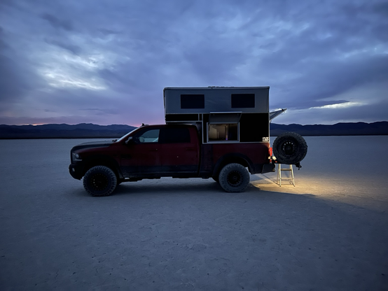 A red RAM Power Wagon with an OVRLND pop-up camper is parked on a dry lake bed at dusk. The camper’s rear hatch is open, and a small step ladder is set up at the back, illuminated by a warm light. Mountains are visible on the horizon under a dark, clouded sky with faint traces of sunset.