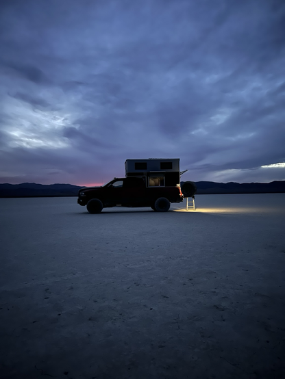 A red RAM Power Wagon with an OVRLND pop-up camper is parked on a dry lake bed at dusk. The camper’s rear hatch is open, and a small step ladder is set up at the back, illuminated by a warm light. Mountains are visible on the horizon under a dark, clouded sky with faint traces of sunset.