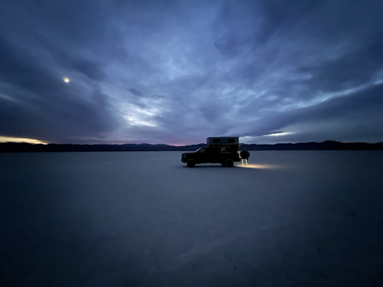 A red RAM Power Wagon with an OVRLND pop-up camper is parked on a dry lake bed at dusk. The camper’s rear hatch is open, and a small step ladder is set up at the back, illuminated by a warm light. Mountains are visible on the horizon under a dark, clouded sky with faint traces of sunset.