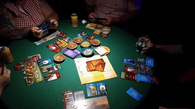 An overhead view of a seven player play of 7 Wonders, players sitting in a circle around the various components spread over a green felt round tabletop