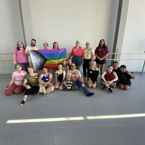18 people, mostly standing, about 6 sitting, pose with a progress pride flag and a sign saying "queer tango collective"