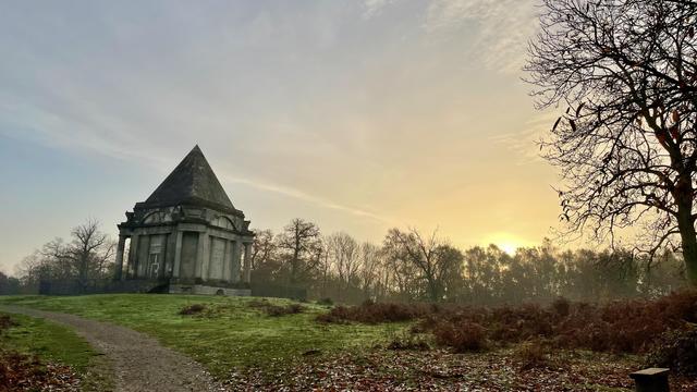 A stone building with a steep pyramid roof stands on a grassy hill at sunrise, framed by bare trees and a cloudy sky.
