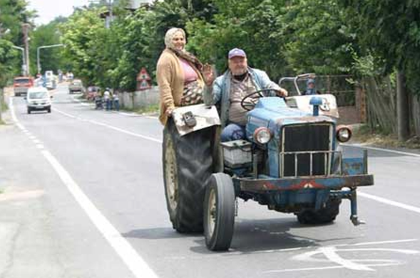 A farming tractor being driven along a street with the front left wheel missing. It's counterbalanced by the farmer's wife (presumably) perched on the rear right wheel guard.