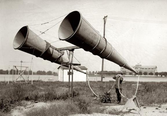 The image depicts a historical scene featuring two large, conical metal structures resembling early radio antennas or acoustic devices, mounted on a metal frame. These structures are positioned on a grassy field, with one slightly larger than the other, both angled towards the sky. A person, dressed in early 20th-century attire, stands near the smaller structure, holding a device that appears to be a microphone or receiver, suggesting an experiment or communication activity. The background includes a small white building, possibly a shed, and a large building with a dome, likely a public or institutional structure. Utility poles and wires are visible, indicating the presence of early electrical infrastructure. The scene is monochromatic, emphasizing the historical context.