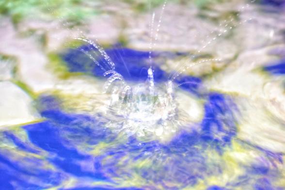 A blurry, overexposed photo of a water drop splashing in a fountain. The central water drop is a bright, luminous white mass with tiny droplets spraying outwards in an ethereal, starburst-like pattern. The background is a soft-focus, abstract blend of blue and yellowish-green, likely reflections from the fountain's tiles. The image has a dreamy, otherworldly quality due to the motion blur and bright light, making the water seem to glow.