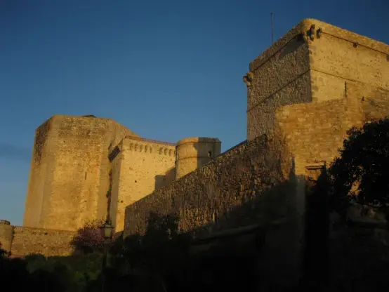The image features a stone castle or fortress structure, likely dating back several centuries. It displays thick stone walls with varying textures and shapes, including towers and battlements. The lighting suggests a time around sunset, casting shadows and highlighting the texture of the stone. Surrounding trees and possibly some vegetation can be seen, adding to the historical and cultural atmosphere of the setting.

Image Credits: Wikimedia / José Manuel Lira Vidal / Public domain