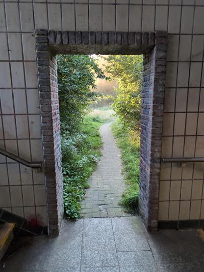 eine Tür ohne Flügel, durch die man von der gefliesten Bahnhofstreppe ins Grüne sehen und gehen kann