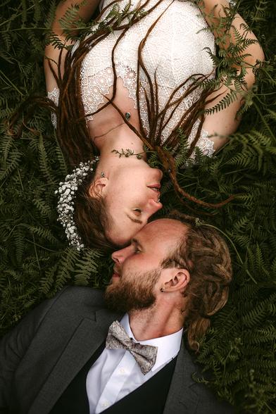 This is a romantic portrait of an elopement in Sweden a couple lying together on a bed of green ferns, photographed from above by European Wedding Photographer Sturmsucht. The woman wears an elegant white lace dress and has long brown hair adorned with small white flowers (baby's breath). The man is dressed in a dark suit with a white shirt and patterned tie, and has brown hair and a beard.

They're positioned with their faces close together in an intimate pose, both lying on their backs against the lush fern backdrop. The woman's hair flows gracefully across the greenery. The soft, natural lighting creates a dreamy, ethereal quality to this wedding or engagement photograph that beautifully blends formal portraiture with organic, forest-like elements.