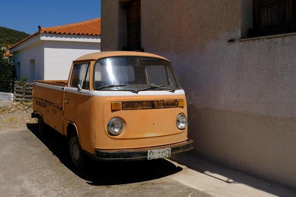 Photo of a matte orange vintage Volkswagen pickup, parked next to a house in a Greek village.