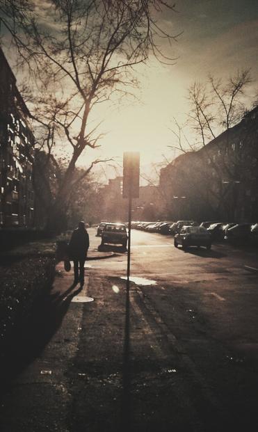 A high-contrast, sepia-toned, vertical photo of a street scene. A person is walking away from the camera on a sidewalk on the left side of the frame. The sun is low in the sky, creating long shadows and a bright lens flare behind a street sign in the center. Parked cars line the street on the right. Bare trees are visible against the sky.