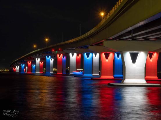 The Ocean City 9th Street Bridge is lit in red, white, and blue lights for the summer season.  The pillars are brightly lit reflecting on the smooth water below.