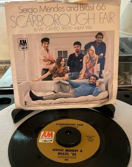 A records sleeve on a stand above a 7" record on a turntable. Sleeve lists at the top in a thin font "Sergio Mendes and Brasil 66" "Scarborough Fair" "B/W Canto Triste A&M 986" The lower ⅔ shows a smiling man laying on a couch, with a group of men and women standing behind it.

The record label is a golden brown, with "Scarborough Fair" in thick letters above the spindle hole. A large A&M Records logo to the left of the spindle hole. Below the hole is "Sergio Mendes & Brasil '66".