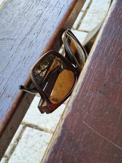 Unas gafas de sol cerradas de pastas negras y cristales marrones. Se encuentran atrapadas entre dos tablas de madera de un banco de parque como si se le hubieran caído a alguien del bolsillo.