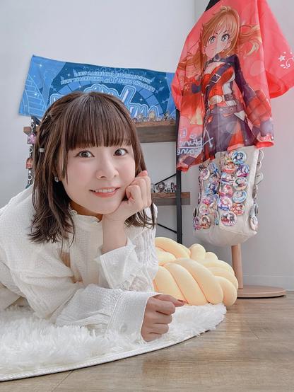 A woman with short brown hair and bangs is lying on a white rug, smiling at the camera. Behind her, a red shirt featuring an anime character and a collection of pins is displayed on a stand, with a blue towel hanging on the wall. The scene has a cozy indoor setting with soft lighting and a relaxed atmosphere.