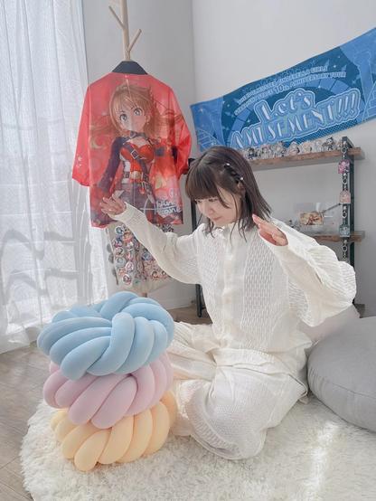 A young woman in a room adorned with anime and manga-inspired decorations. The woman has brown hair, which is styled in a braid on the right side of her head. She wears a white, long-sleeved blouse and white pants, and is seated on a white shag rug. Her hands are raised in front of her, and her palms are facing each other.
To the left of the woman is a colorful, knotted pillow with pastel hues of pink, blue, and yellow. Behind her, on the right, is a shelf displaying a collection of figurines. The wall behind the woman features a blue banner with white text, which reads "Let's Amusement!!" in a playful, cursive font. Above the banner is a red shirt with an anime character printed on it, hanging from a coat rack.
The room's walls are painted white, and the floor is made of light-colored wood. The overall atmosphere of the image is one of coziness and comfort, with the woman's relaxed pose and the soft, pastel colors of the room creating a sense of warmth and tranquility.