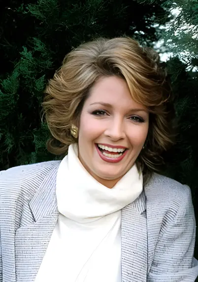 Deidre Hall in 1984, smiling brightly in a striped blazer and white turtleneck, framed by lush greenery—radiating joy, timeless style, and the effortless charisma that defined an era.