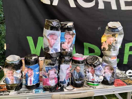 Auf einem Tisch stehen Getränkedosen, auf die Bilder von rechten Politiker*innen geklebt sind. Sie sind sehr verbeult.
