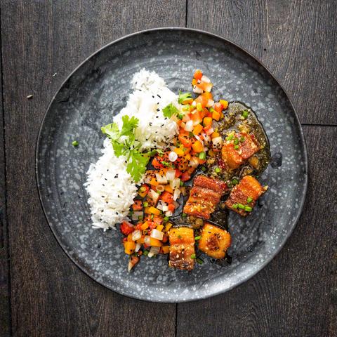Auf einem dunklen Teller befinden sich zwei Nocken Gesichter Reis, ein Streifen mit gleichmäßig gewürfelten Mix aus Kürbis, Wassermelone und Rettich, durchsetzt mit Lauchzwiebeln,  und 5 Würfel knusprig braun gebratener Schweinebauch in einer mit Ingwer und Chili gewürzten Karamellsauce