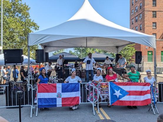 A photo of the salsa band The Ray Delgado Project performing at a street festival in New York City. The band includes keyboards, upright bass, multiple percussionists, vibraphone and a lead singer. There is a line of women dancers in front of the stage. There are flags of Puerto Rico and the Dominican Republic hanging on the barriers in front of the dancers.