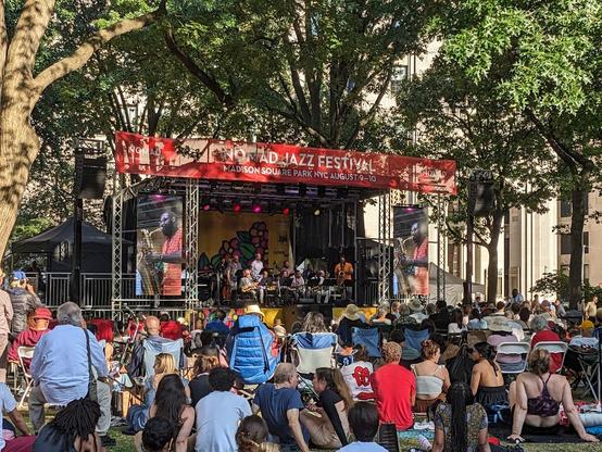 Photo of the Roy Hargrove Big Band on stage at the Nomad Jazz Festival in NYC's Madison Square Park. There are large screens on both sides of the stage showing a shot of baritone saxophonist Jason Marshall playing a solo. Spectators are sitting and standing in the park all around the stage.