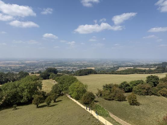 A panoramic view of a lush green landscape with rolling hills, lined paths, scattered trees, and distant villages under a clear blue sky with a few clouds.
