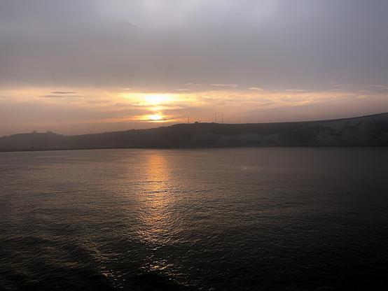 Sunset over a calm sea with golden light reflecting on the water. In the background, hilly terrain with low-lying buildings and a few visible antennas silhouetted against the dimming sky.