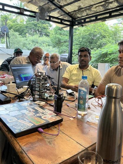 Group of people around a talk with various things lying on the table including laptop, bottles etc.
