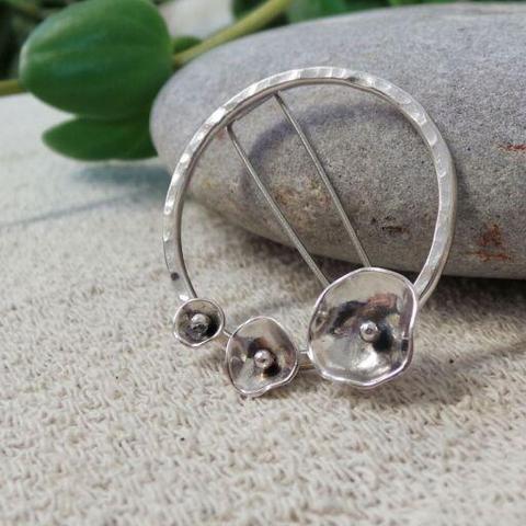 A brooch made of a textured silver ring with three silver blooms attached. It is resting against a grey stone. Some green plant leaves are in the background.