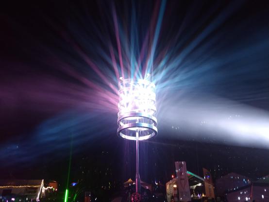 A towering column of stacked mirrored rings catches a brilliant core of white light, bursting it into radiant beams of purple, blue, and silver that streak across the night sky; below, a vibrant patchwork of tents and festival stalls shimmers in the glow.