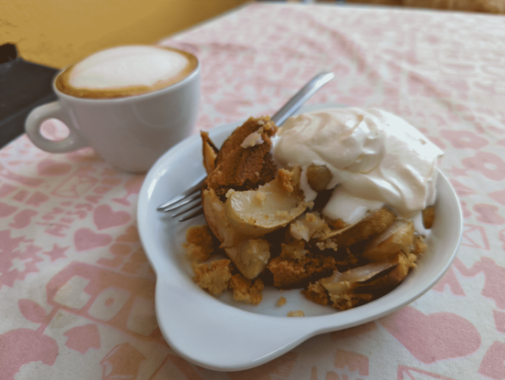 Ein leicht lädiertes Stück Apfelkuchen mit einem großzügigen Schlag Sahne. Eine Tasse mit Cappuccino steht daneben, auf einem Tisch mit ausgeblichen-gemusterter Tischdecke neben einer sonnenbeschiedenen gelbem Wand.