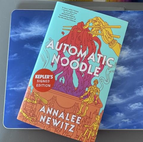 A hardcover copy of the book Automatic Noodle by Annalee Newitz, sporting a red sticker that says “Kepler’s Signed Edition”. The brightly colored book cover depicts a robotic hand using chopsticks to hold a bundle of big noodles. Below that are sketched outlines of scenes and objects from the story, including three of the robot characters and the Golden Gate Bridge.

The laptop below the book sports a skin with a photograph I took of wispy cirrus clouds against a deep blue sky.