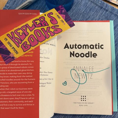 A copy of Automatic Noodle by Analee Newitz open to the title page, where it is autographed in teal ink with a tiny heart and a looping line that might be a loose signature or a noodle, or both. Atop the open book is bookmark that says Kepler’s Books, Menlo Park, CA, established 1955.