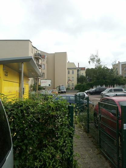 Ein Stück überwucherter Zaun, Ausschnitt einer Packstation. Autos.