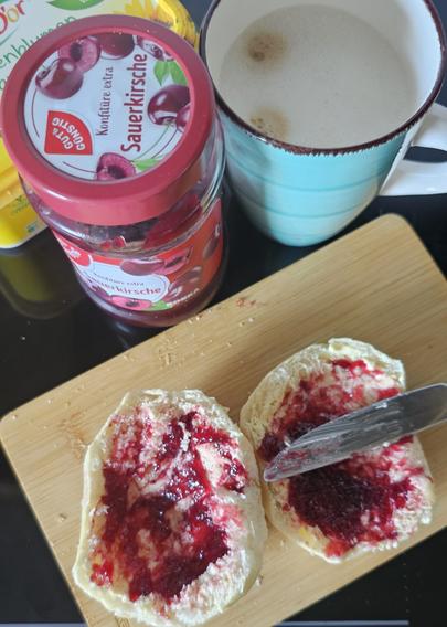 Das Bild zeigt eine appetitliche Mahlzeit auf einem dunklen Tisch. Im Vordergrund befindet sich ein Holz-Schneidebrett, auf dem zwei Butterbrote liegen, die mit roter Kirsch Konfitüre bestrichen werden. Ein Messer mit scharfen Kanten ist in der Konfitüre eingetaucht und wird verwendet, um sie auf die Brote zu verteilen. Neben dem Schneidebrett steht eine grüne Tasse mit einem cremigen Getränk, das möglicherweise Kaffee oder Tee ist. Im Hintergrund ist ein Glas mit roter Kirschkonfitüre zu sehen, das mit dem Etikett "GUT & GÜNSTIG Sauerkirsche Konfitüre extra" gekennzeichnet ist. Die Konfitüre ist in einem transparenten Glas mit einem roten Deckel verpackt und zeigt Bilder von Kirschen auf dem Etikett. Die gesamte Szene vermittelt ein Gefühl von gemütlicher Morgenstimmung.