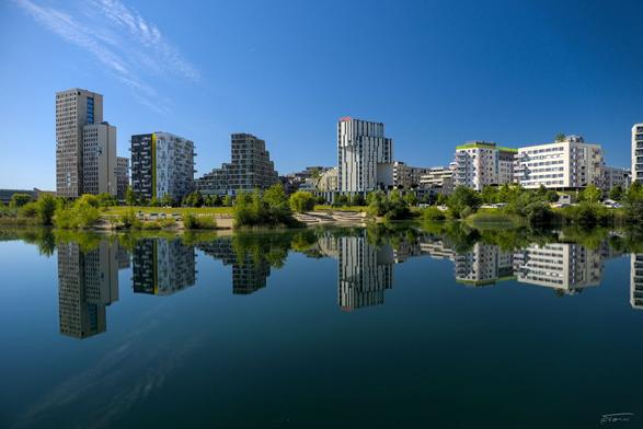 Das Bild zeigt eine moderne Stadtlandschaft, die sich am Ufer eines ruhigen Gewässers befindet. Die Skyline besteht aus mehreren hohen Gebäuden mit zeitgenössischer Architektur, darunter Wohn- und Bürogebäude. Die Gebäude spiegeln sich klar im Wasser, was eine symmetrische und ästhetisch ansprechende Szene schafft. Im Vordergrund gibt es grün bewachsene Flächen mit Bäumen und Büschen, die einen Kontrast zu den urbanen Strukturen bilden. Der Himmel ist klar und blau. Die Szene wirkt friedlich und gut geplant, typisch für eine moderne Stadtentwicklung wie die Seestadt Aspern.