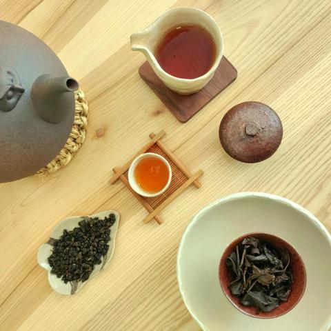 A gong fu cha set seen from above: a clay kettle on the left, next to which is a ceramic tea scoop with tightly rolled tea leaves, a small white cup on a wooden coaster, a ceramic pitcher, and finally a dark gaiwan full of opened, quite dark oolong leaves.