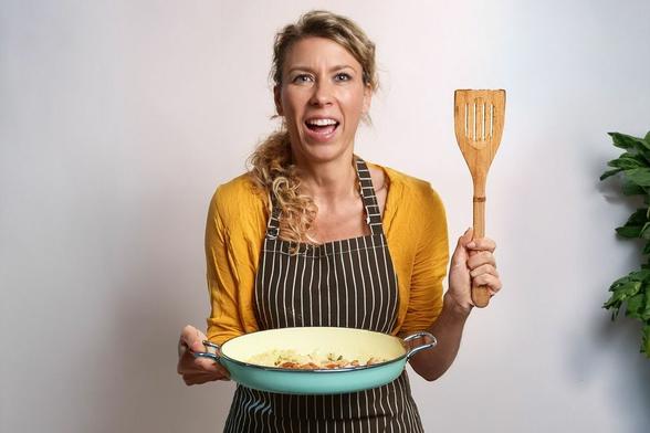 Una mujer con una camisa amarilla y un delantal de rayas sostiene una sartén con pasta y verduras en una mano y una espátula de madera en la otra. Sonríe y está sorprendida porque acaba de transformar un error culinario en un plato nuevo.