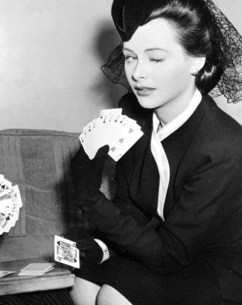 Publicity photo of Hedy Lamarr. She is a white woman with darkish hair, playing cards and theatrically winking to camera as she shows the audience her hand.