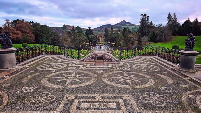 A wide shot of the intricate pebble and stone patio at Powerscourt Gardens, Ireland, with the year 1875 formed in the stones at the bottom of the image. The ornate terrace features a detailed compass rose design and a decorative metal railing with gold accents. Beyond the terrace, a lush green lawn leads to a forest of trees, with a large, distinctive mountain visible in the distance under a cloudy evening sky. Two classical statues on pedestals frame the foreground on either side of the terrace.