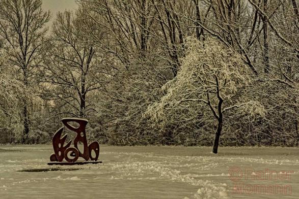 Farbfotografie im Format 3:2 Landscape.
Winterbild eines verschneiten Parks. Restlicht und Straßenbeleuchtung geben die Lichtmischung zu dieser Langzeitbelichtung.
Die rostige Skulptur "Les Plantes Plasmatiques 1" steht der Schnee bedeckten, Wiese. Links, im unteren Bereich. Rechts ein zweieinhalbfach größerer Baum, mit schneeweißen Ästen, welche sich nach der Skulptur strecken. Beide bilden eine harmonische Einheit. Im Hintergrund mehrere große Bäume, die über die Fotografie hinausragen.
Oval, Kreis, Kelch, Blatt und Blüte – weich und organisch wie einen Naturwerkstoff versteht der Künstler den rostroten Cortenstahl zu einer abstrakten, sanftlinigen Skulptur zu formen.

https://www.misterhex.de/