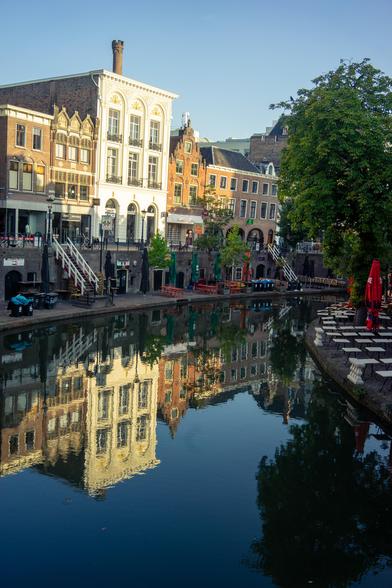 This image depicts a serene canal scene in what appears to be a European city, likely one known for its picturesque waterways and historic architecture. The canal is flanked by an array of colorful buildings on one side, reflecting in the calm water. The area is adorned with outdoor seating, trees, and a clear blue sky overhead.

On the left side, various buildings of different architectural styles stand in a row. Prominently, there's a larger, light-colored building with tall windows and ornate detailing, surrounded by smaller, similarly styled buildings featuring dark, stepped-gable roofs and warm-toned facades. The street level is adorned with cafes, covered outdoor seating areas, and small trees planted along the canal's edge, contributing to a welcoming ambiance. The water in the canal is still, providing a mirror-like reflection of the buildings and trees. The clarity of the reflections suggests the water is clean and undisturbed. On the right side, there is a set-up of outdoor café tables and chairs, with umbrellas and greenery enhancing the tranquil environment. A tree with lush green foliage is also visible, adding to the scene's serenity. The overall setting appears to be calm and inviting, likely a popular spot for locals and tourists to enjoy the scenic view.