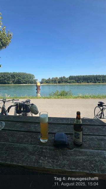 Ein malerischer Blick auf einen Fluss mit üppigem Grün im Hintergrund. Im Vordergrund stehen zwei Fahrräder neben einem Holztisch. Ein Glas Bier und eine Flasche stehen auf dem Tisch, mit einem klaren blauen Himmel darüber.