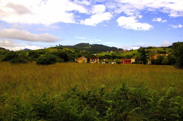 Un campo de hermosa yerba con unas pocas casas unifamiliares detrás, al fondo un monte bajo un cielo azul acompañado de diferentes nubes con personalidad propia.
