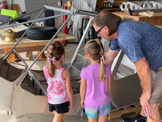 Two young girls inspecting a partially build airplane while an adult explains what they're looking at.