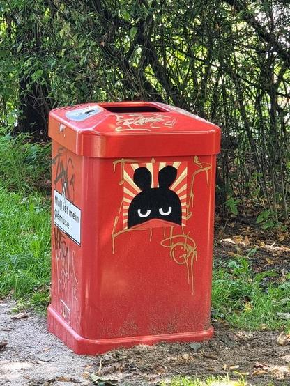 Foto eines feuerroten zylinderförmigen öffentlichen Mülleimers, der mit einem großen rechteckigen Sticker beklebt ist, welcher eine mit einem roten Tuch maskierte silhouettenartig schwarze Maus zeigt, und zwar diese Maus aus der Sendung mit der Maus mit ihrem eiförmigen Körper und den aufgestellten Ohren. Allerdings ist die Maus aus der Sendung orange, und die auf dem Sticker ist flächig schwarz und ihre Augen nicht etwa typisch liebevoll entspannt, sondern etwas wütend oder entschlossen. Der Hintergrund der Maus besteht aus fächerförmig auseinandergehenden gelb-roten Streifen, die wie stilisierte Strahlen einer hinter ihr stehenden Sonne wirken. Um den Mülleimer herum ist grüne Wiese, sandiger Weg und Blätterwerk von Bäumen.

Nachtrag: Auf der Seite des Mülleimers klebt eine Sprechblase mit dem Text "Müll ist mein Gemüse". Auf diesen und andere typische Sprüche auf Hamburger Mülleimern beziehen sich viele Kommentare zum Beitrag. Ich selbst habe diesen Spruch gar nicht bemerkt und ergänze ihn deshalb erst jetzt hier im AltText.