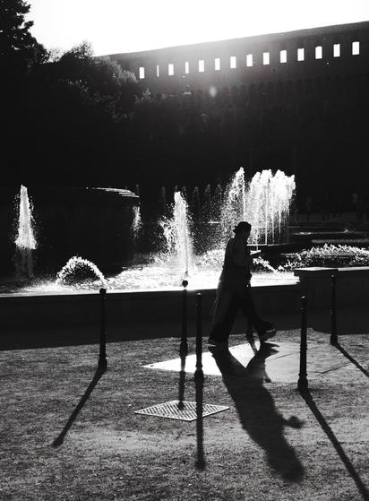 A woman walks past a fountain with strong water jets. The backlit scene has stark contrast between light and shadow, and the woman projects a long shadow towards us, while her silhouette is visible againts the bright water jets.